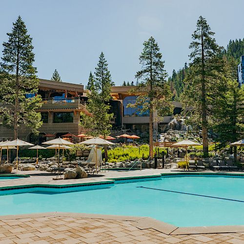 A resort with a large swimming pool, surrounded by lounge chairs and umbrellas, set amidst trees and buildings.