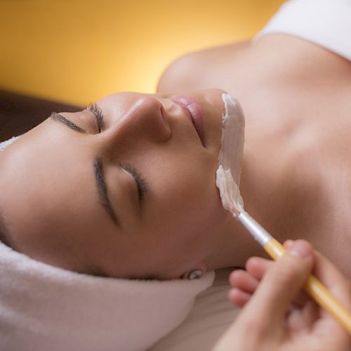 A person is lying down with a towel wrapped around their head, receiving a facial treatment involving a brush applying a substance to their face.