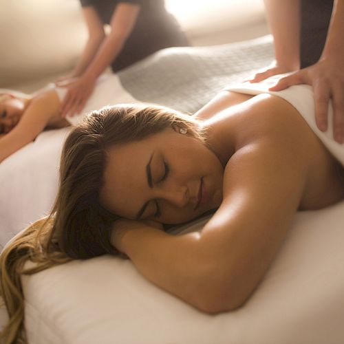 Two people are receiving massages on massage tables, with therapists working on their backs in a relaxing setting.