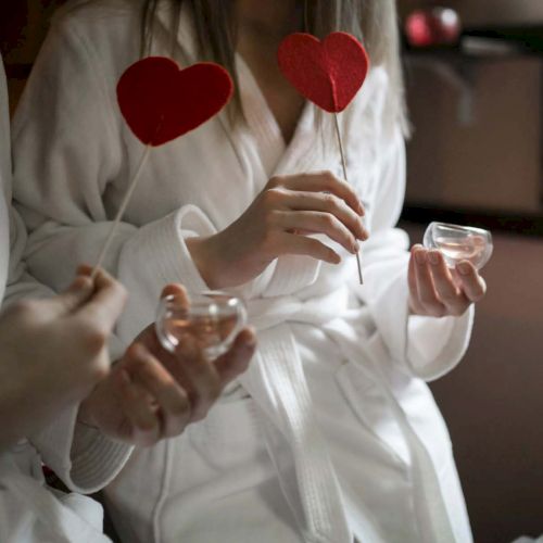 Two people in white robes hold heart-shaped decorations and small cups, suggesting a romantic or relaxing setting.