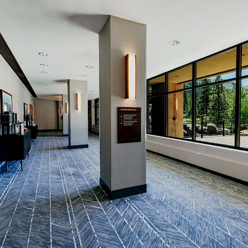 A hallway with wooden doors, framed pictures, large windows showcasing a scenic view, and a central pillar with a sign is pictured.