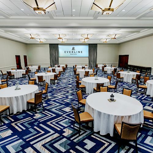 An event hall with round tables covered with white tablecloths and surrounded by chairs. A screen at the front displays 