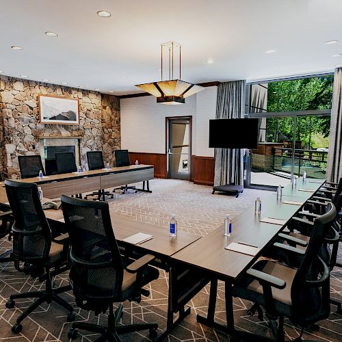 A modern conference room with a large table, several office chairs, water bottles, notepads, a TV screen, large windows with curtains, and a stone wall.