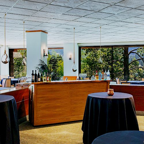 A well-lit banquet or reception area with round tables clothed in black, candles, and a wooden bar setup with chairs and bottles, in a room with large windows.