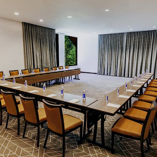 A conference room with a U-shaped table setup, chairs, notepads, pens, and water bottles on the table; large windows with curtains.