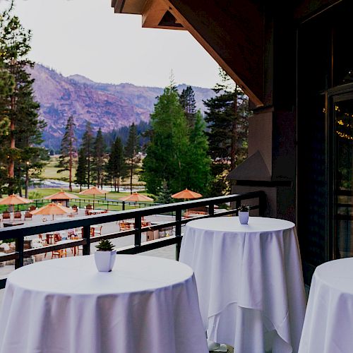 A scenic view from a balcony with round tables covered in white cloths and small potted plants, overlooking a mountain landscape and outdoor seating.