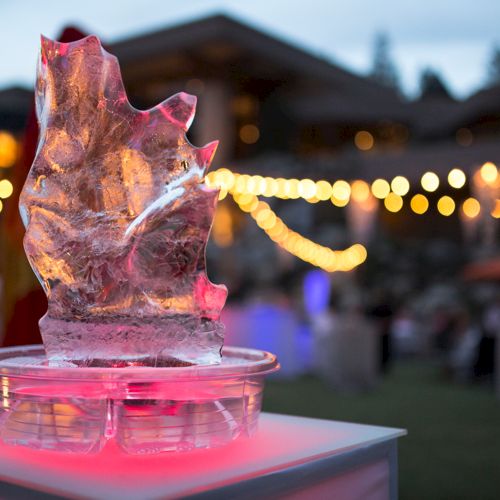 A lit, colorful ice sculpture shaped like an abstract form sits on a table at an outdoor event with glowing lights behind.