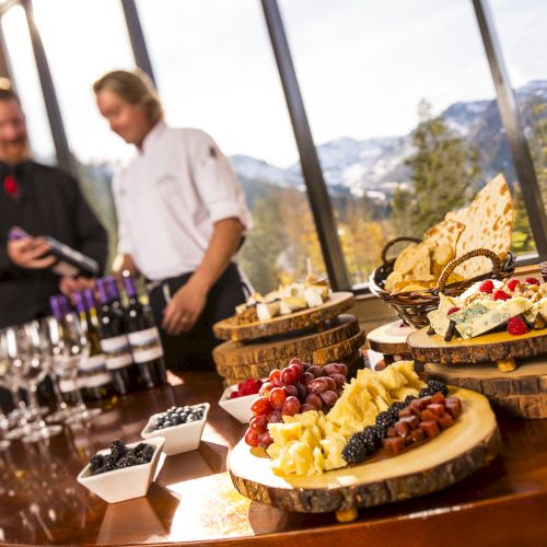 A wine-and-cheese tasting spread with sliced cheese, fruits, and grapes by a window as two servers pour with a mountain view.