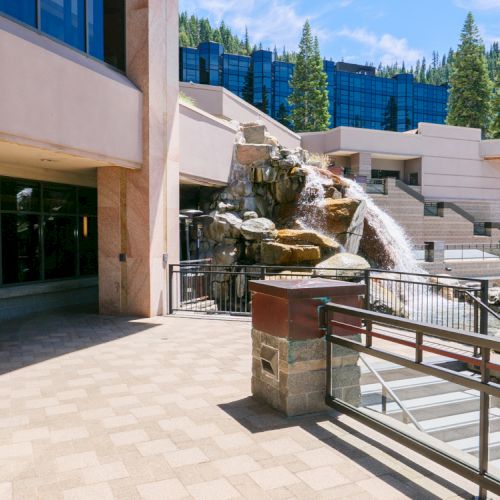 A modern hotel courtyard with a stone water fountain and pine trees in the background.