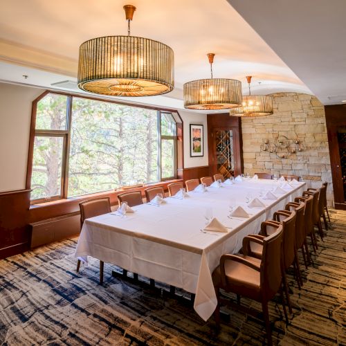 A fine-dining room with a set table, pendant chandeliers, large windows, and a stone wall with a wine display.