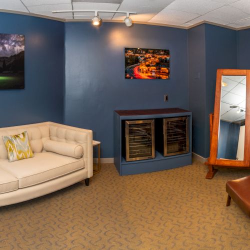 A cozy lounge with a white sofa, wooden chairs, framed photos, two wine fridges, and a wall mirror under ceiling lights.