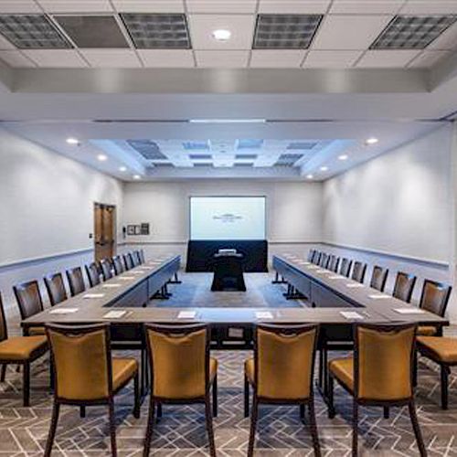 A modern conference room with a U-shaped table setup, chairs around it, and a projector screen at the front. The room is well-lit with ceiling lights.