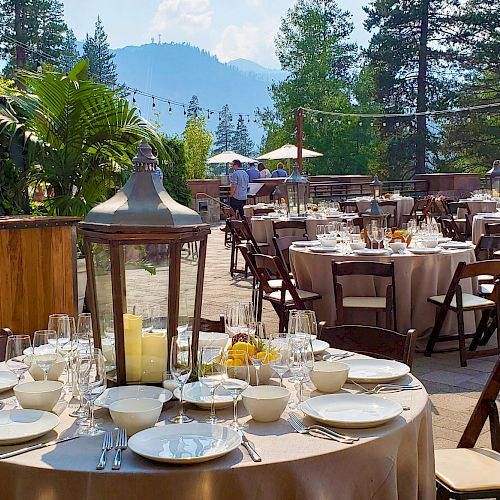 The image shows an outdoor event setup with round tables adorned with tableware and centerpieces, surrounded by scenic mountains and trees.