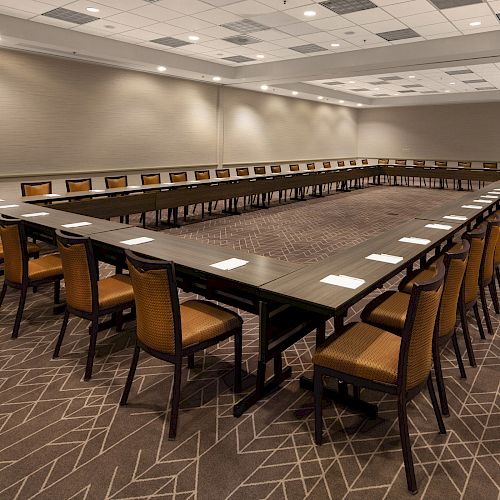 A conference room with U-shaped table setup, surrounded by chairs; notepads are placed on the table, and the room has carpeted flooring and ceiling lights.