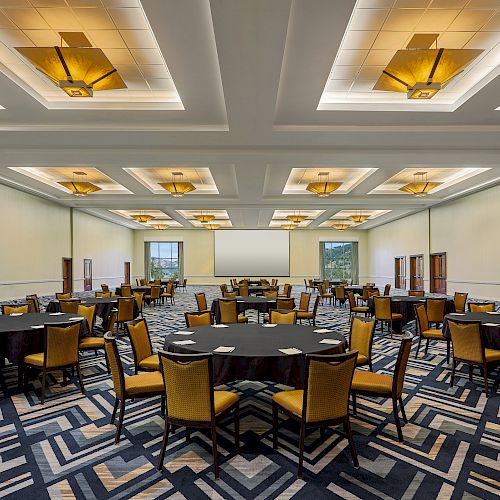 The image shows a spacious conference room with round tables, yellow chairs, patterned carpet, rectangular lighting fixtures on the ceiling, and a projector screen.