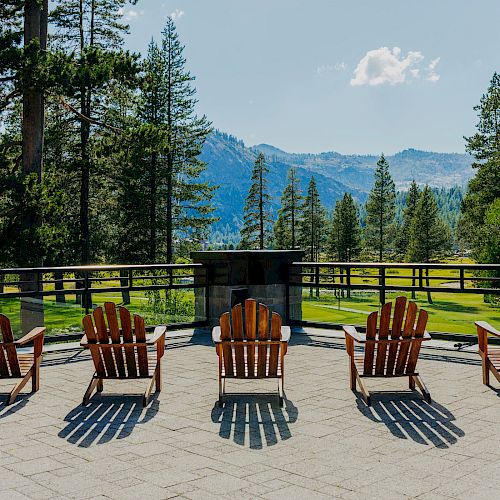 Four wooden chairs face a scenic view of mountains and trees on a sunny day, with a railing and paved patio in the foreground.