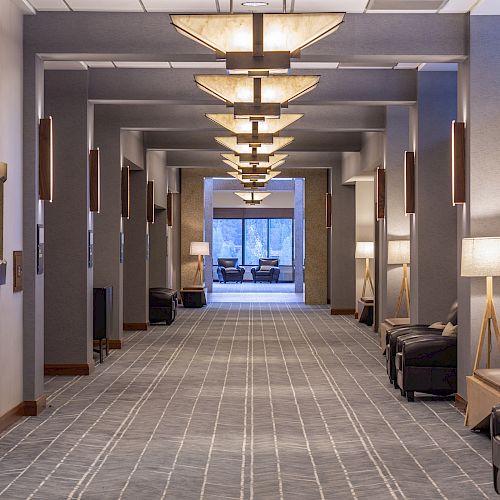 A hallway with carpeted floors, modern ceiling lights, side lamps, and seating areas, leading to a room with large windows.