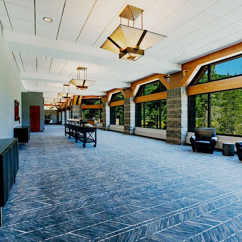 A spacious lobby with large windows showcasing a green, wooded view. It has modern lighting fixtures, a blue-patterned carpet, and several seating areas.