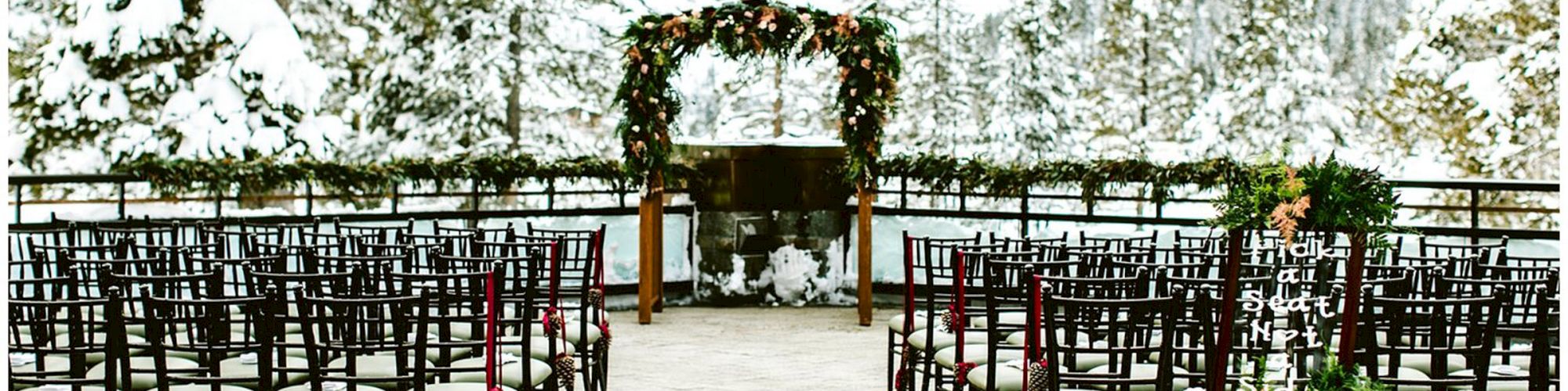 An outdoor wedding set up with chairs aligned facing an arch, set in a snowy landscape with mountains and trees in the background.