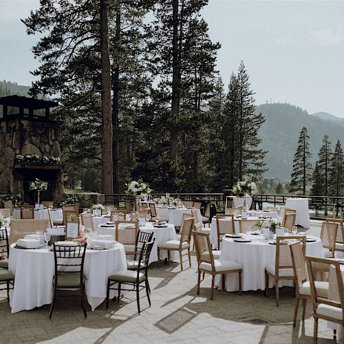 This image shows an outdoor event setup with round tables and chairs in a scenic, forested mountain area, decorated with floral centerpieces.