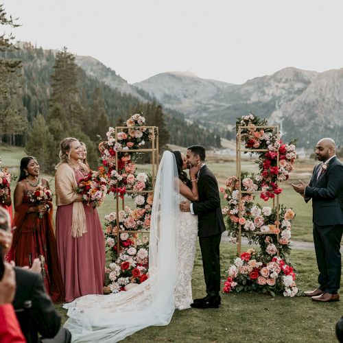 An outdoor wedding ceremony with a bride and groom kissing, surrounded by floral pillars, applauding guests, and mountains in the background.