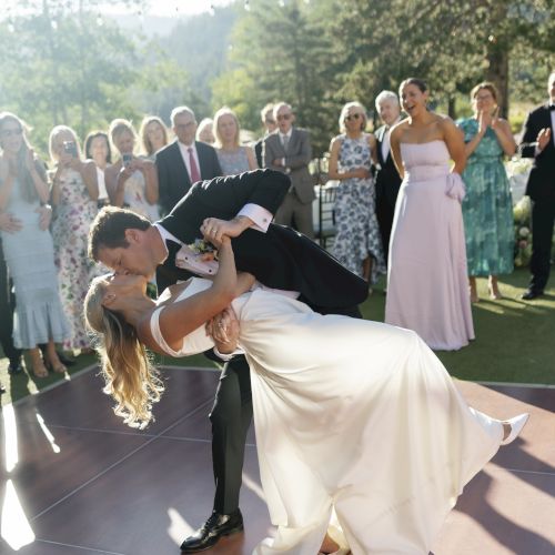 An outdoor wedding moment: the groom lifts the bride in front of guests, string lights overhead, celebrating their big day with joy.