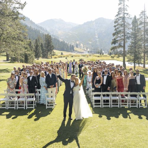 A large wedding party stands on a grassy outdoor aisle in the mountains, waving as the bride and groom celebrate.