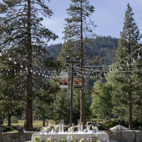 Outdoor wedding reception setup with a flower-covered table, string lights hanging between tall trees, and mountains in the background.