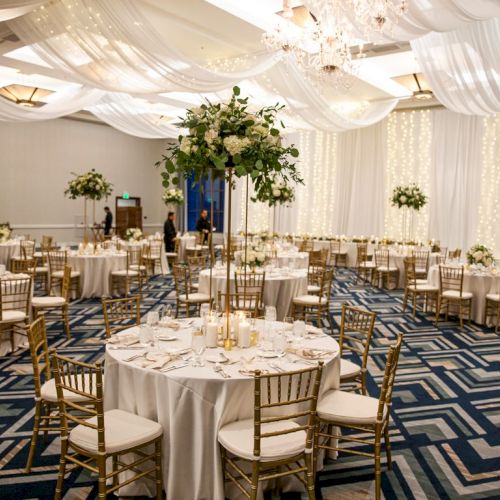 An elegant wedding reception hall with candlelit tables, gold chairs, floral centerpieces, white drapes, and crystal chandeliers.