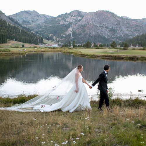 A bride and groom in wedding attire walk hand-in-hand by a calm lake, with alpine mountains in the background.