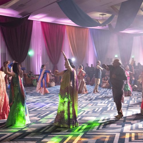 A lively dance floor at a cultural event, with people in colorful traditional outfits dancing under pink and green stage lights.