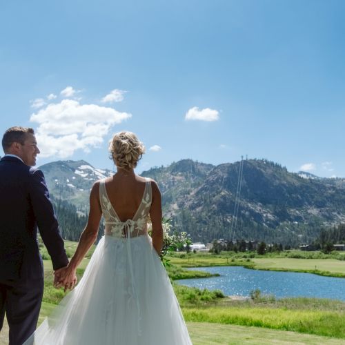 A bride and groom hold hands outdoors at a wedding by a pond, with mountain views and blue sky behind them.