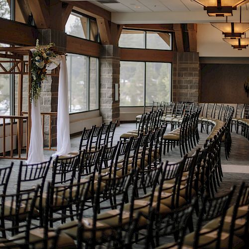 It's an empty indoor wedding ceremony setup with rows of chairs and a decorated arch near large windows.