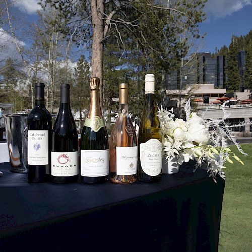 A table outdoors with five wine bottles, a wine cooler, and floral arrangements. Trees and buildings are in the background under a blue sky.