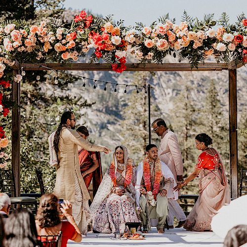 An outdoor wedding ceremony under a decorated floral arch with the couple seated and family members standing around them in traditional attire.