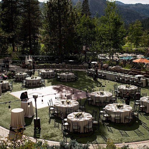 An outdoor event setup with round tables, chairs, and decorations on a green lawn surrounded by trees and mountains in the background.