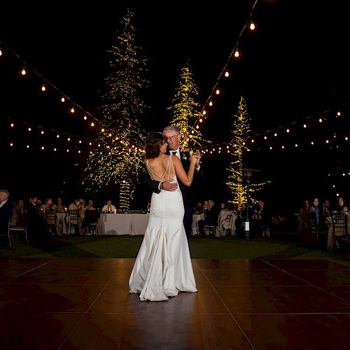 A couple dances under string lights at an outdoor wedding reception, surrounded by guests seated at tables during the night.