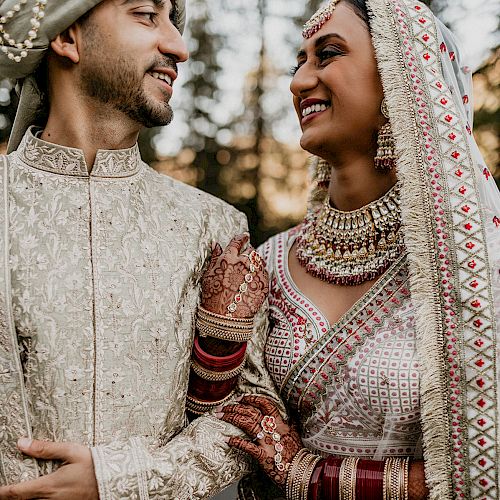 A couple dressed in traditional wedding attire smiles at each other outdoors. They are wearing intricately designed outfits adorned with jewelry and henna.