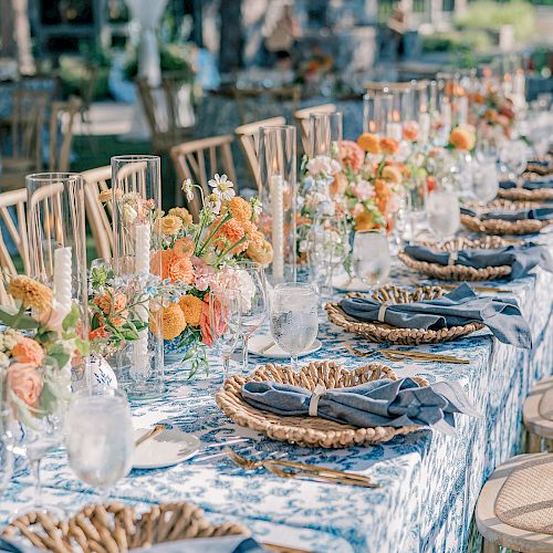 An elegantly set outdoor dining table with floral centerpieces, wicker placemats, light blue napkins, candles, and chairs, ready for a special event.