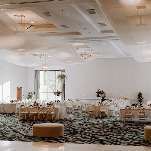 The image features a spacious, elegantly decorated event hall with round tables, chairs, floral arrangements, and draped ceiling fabrics.