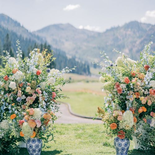 Outdoor wedding aisle with colorful flower vases on both sides, set against green mountains and a cloudy sky.