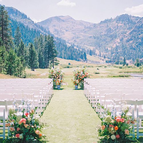 An outdoor wedding setup with white chairs arranged on either side of an aisle adorned with colorful flower arrangements, against a mountain backdrop.