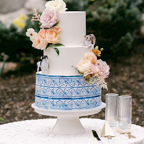 A three-tiered white cake decorated with flowers and blue patterns is on a stand with glasses and cutlery on a white tablecloth.