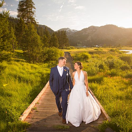 A couple in wedding attire walks hand-in-hand on a wooden path through a lush green meadow with mountains and a lake in the background during sunset.