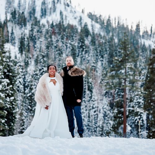 A couple stands in the snow with a mountain and forest backdrop. The woman wears a white dress, and the man wears dark clothing with a fur collar.