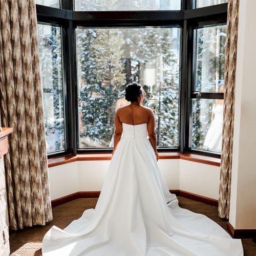 A bride in a flowing white gown stands before a large window, gazing outside. The room has curtains and sunlight pours in, highlighting her dress.