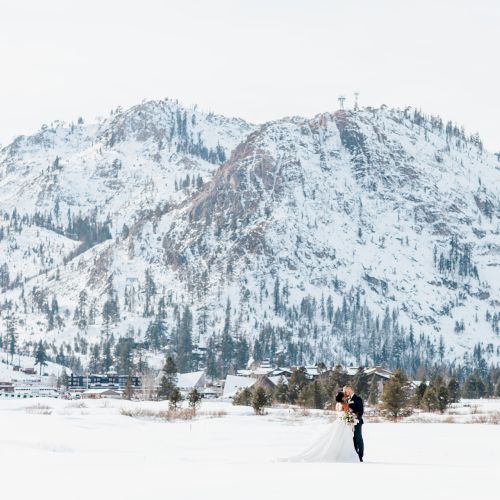 A bride and groom stand in a snowy landscape with mountains in the background, creating a picturesque winter wedding scene.