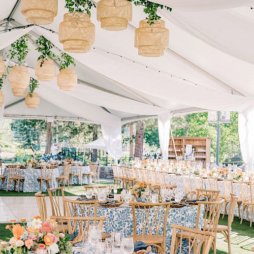 The image shows a beautifully decorated outdoor event setup with tables, chairs, floral arrangements, and hanging lanterns under a white canopy.