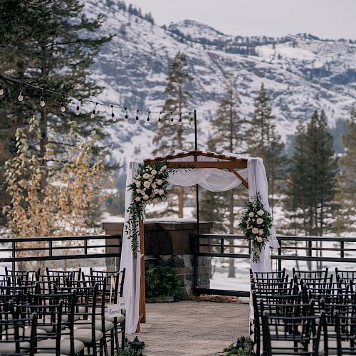 An outdoor wedding setup with rows of chairs facing a floral arch, framed by snowy mountain scenery and pine trees in the background.