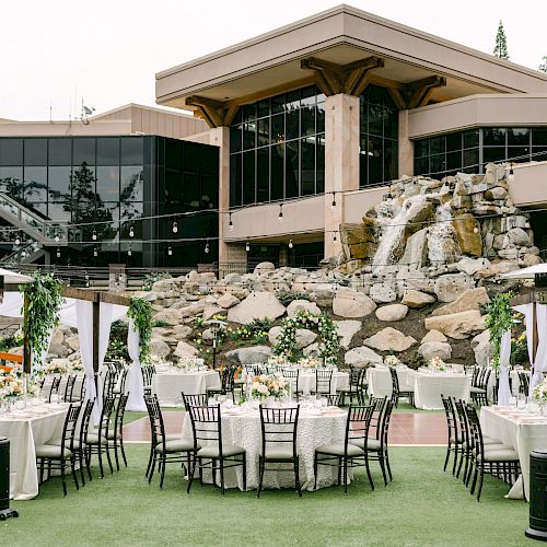 An elegant outdoor event setup with round tables, decorated with white linens and flowers, in front of a building and waterfall.